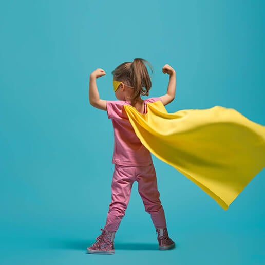 Ein kleines Mädchen in einem Superhelden-Kostüm steht mit dem Rücken zur Kamera. Sie trägt ein pinkes Outfit, eine gelbe Augenmaske und einen flatternden gelben Umhang. Sie hält ihre Arme angewinkelt und zeigt stolz ihre 'Muskeln'. Der Hintergrund ist einfarbig türkis, wodurch das Kostüm und ihre Pose im Vordergrund hervorgehoben werden.